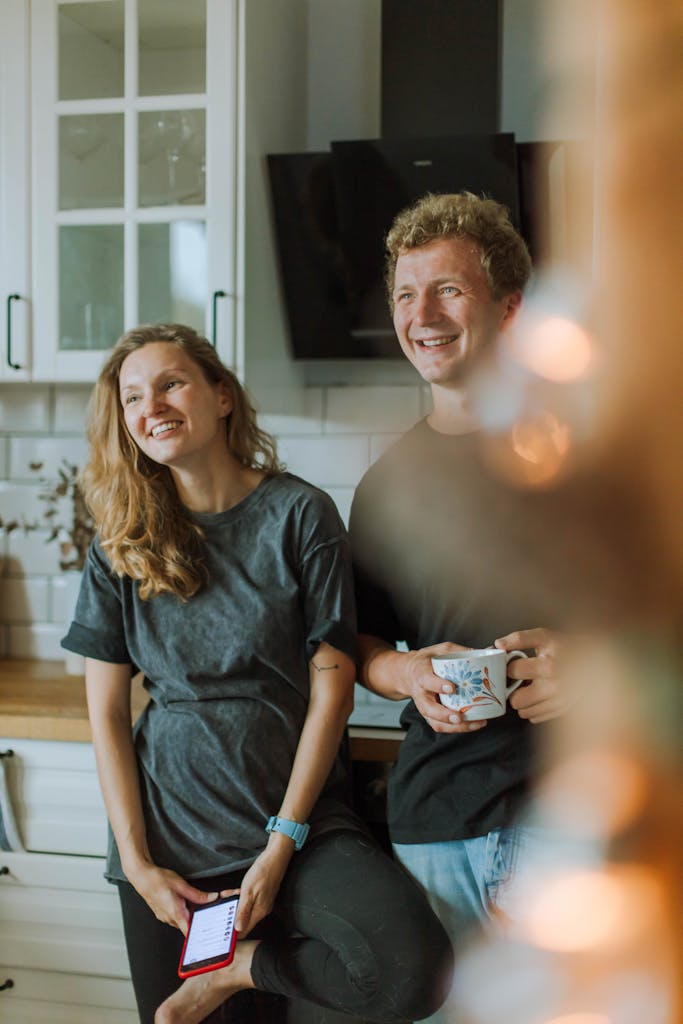 Smiling couple standing in a modern kitchen, enjoying leisure time with a cup and smartphone.